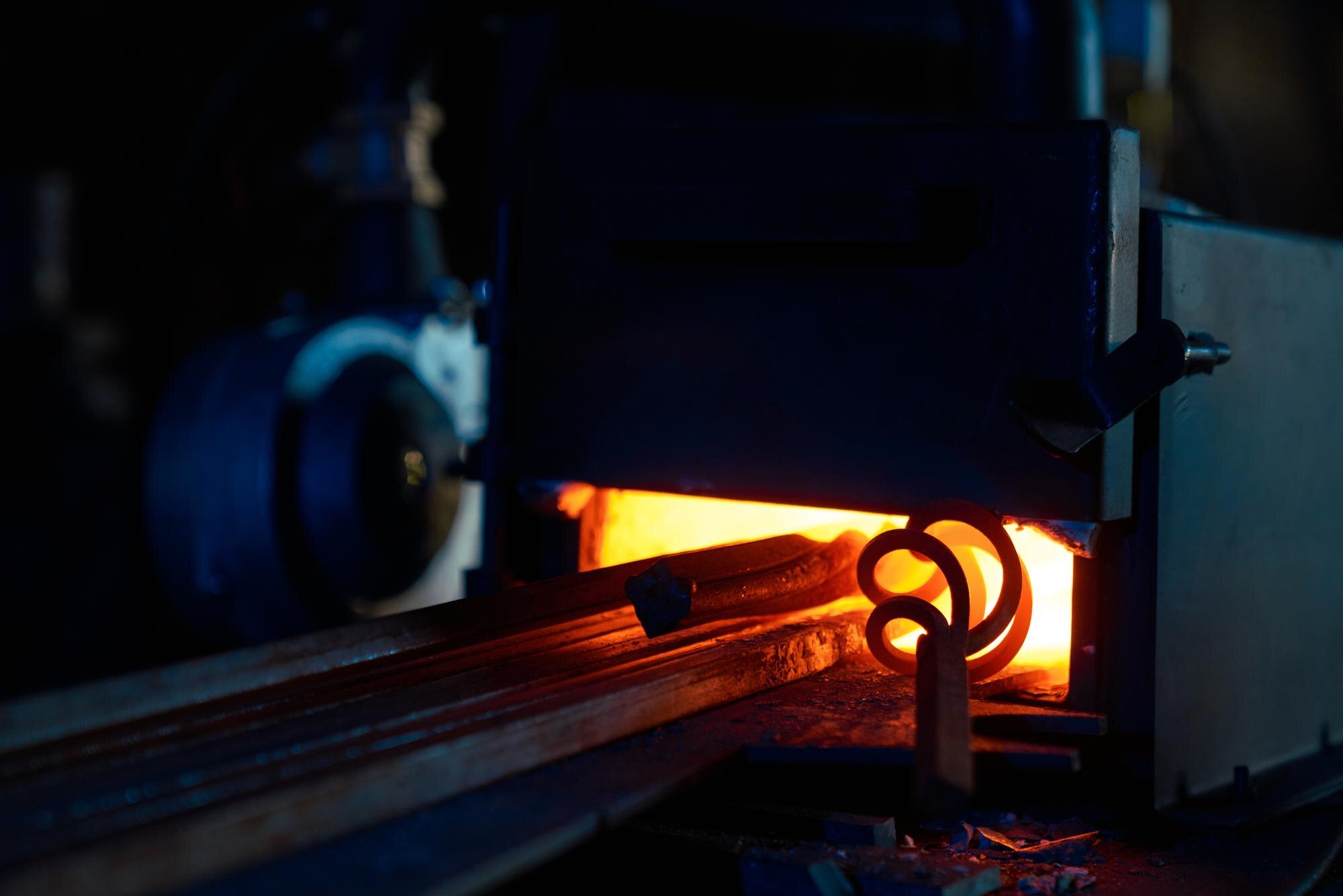 Peças metálicas sendo aquecidas em forno industrial durante o processo de fundição de precisão, com brilho intenso do metal incandescente e ambiente fabril ao fundo.